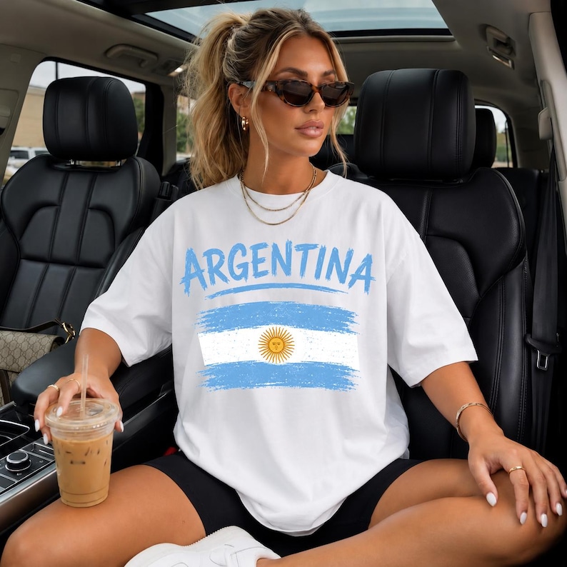 May include: A white short-sleeved t-shirt featuring the word ARGENTINA in light blue letters above a distressed graphic of the Argentine flag, which includes a central yellow sun emblem on horizontal light blue and white stripes.