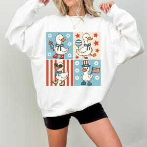 May include: White sweatshirt with a graphic of four cartoon ducks wearing patriotic themed outfits. The ducks are wearing red, white, and blue clothing and accessories. The graphic is set against a background of red and blue stripes, white stars, and white flowers.