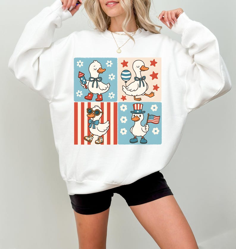 May include: White sweatshirt with a graphic of four cartoon ducks wearing patriotic themed outfits. The ducks are wearing red, white, and blue clothing and accessories. The graphic is set against a background of red and blue stripes, white stars, and white flowers.