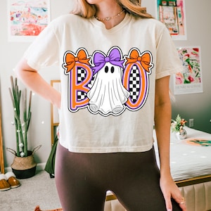 May include: A cream-coloured t-shirt with a Halloween graphic. The word "BOO" is displayed in orange and purple, with a ghost character in the centre. Orange bows decorate the letters, and the background has a chequered pattern.