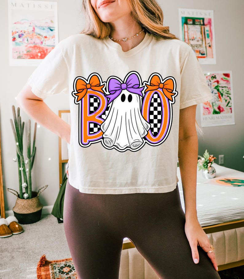 May include: A cream-coloured t-shirt with a Halloween graphic. The word "BOO" is displayed in orange and purple, with a ghost character in the centre. Orange bows decorate the letters, and the background has a chequered pattern.