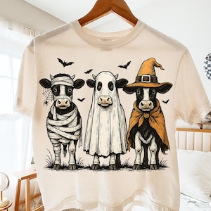 May include: Cream-colored t-shirt featuring three cows dressed in Halloween costumes. One cow is wrapped as a mummy, another is a ghost, and the third is a witch. Bats and a spiderweb are also included in the design.