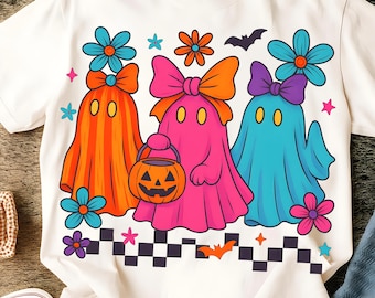 Può includere: T-shirt bianco con un design colorato di Halloween. Tre fantasmi dei cartoni animati in arancione, rosa e blu con fiocchi e fiori. Il fantasma rosa tiene in mano un secchio a forma di zucca. Un pipistrello e delle stelle completano il design.