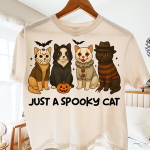 May include: Cream-colored t-shirt featuring a graphic of four cats dressed in Halloween costumes. The cats are wearing costumes of a hockey mask, a vampire, a clown, and a striped sweater. The text "JUST A SPOOKY CAT" is printed below the cats.