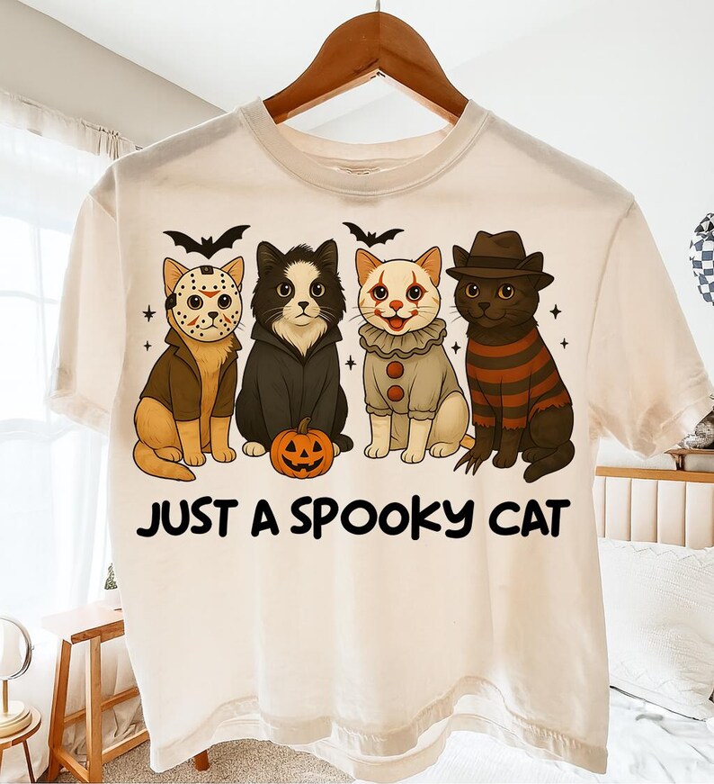 May include: Cream-colored t-shirt featuring a graphic of four cats dressed in Halloween costumes. The cats are wearing costumes of a hockey mask, a vampire, a clown, and a striped sweater. The text "JUST A SPOOKY CAT" is printed below the cats.