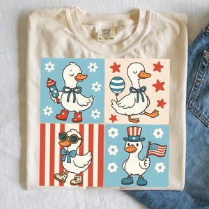May include: A white t-shirt with a design of four cartoon ducks wearing patriotic outfits. The ducks are wearing red, white, and blue clothing and accessories. The design is on a cream background with red and white stripes and blue and white stars.