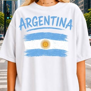 May include: A white short-sleeved t-shirt featuring the word ARGENTINA in a light blue brushstroke font above a graphic of the Argentine flag, which includes horizontal light blue and white stripes and a central yellow sun emblem.