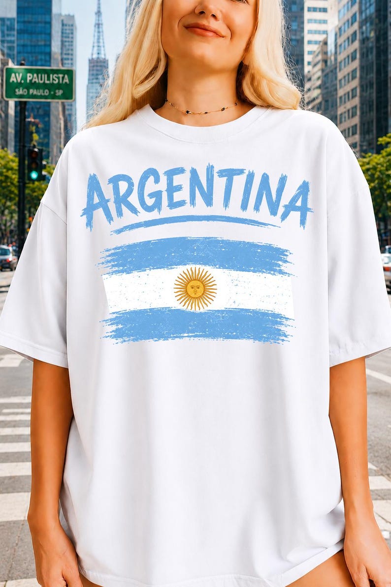 May include: A white short-sleeved t-shirt featuring the word ARGENTINA in a light blue brushstroke font above a graphic of the Argentine flag, which includes horizontal light blue and white stripes and a central yellow sun emblem.