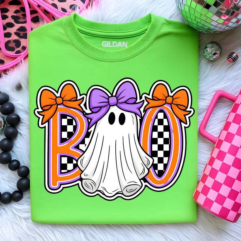 May include: A lime green t-shirt featuring the word "BOO" in orange and purple, with orange bows and checkered patterns. A white ghost with a purple bow is in the centre. The brand name "GILDAN" is printed on the shirt.