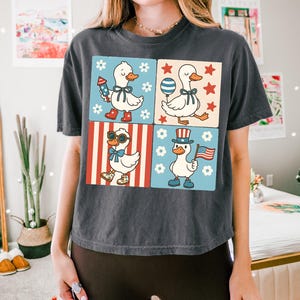 May include: A dark gray t-shirt with a graphic of four cartoon ducks in a patriotic theme. The ducks are wearing red, white, and blue accessories and are surrounded by stars, stripes, and flowers.