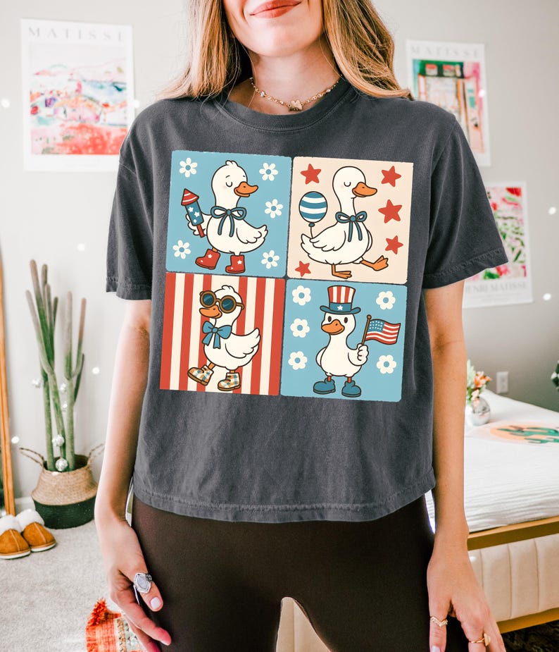 May include: A dark gray t-shirt with a graphic of four cartoon ducks in a patriotic theme. The ducks are wearing red, white, and blue accessories and are surrounded by stars, stripes, and flowers.