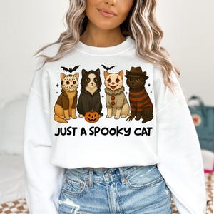 May include: White sweatshirt featuring four cartoon cats dressed in Halloween costumes. The cats include a Jason Voorhees cat, a black cat, a clown cat, and a Freddy Krueger cat. The text "JUST A SPOOKY CAT" is printed below the cats.