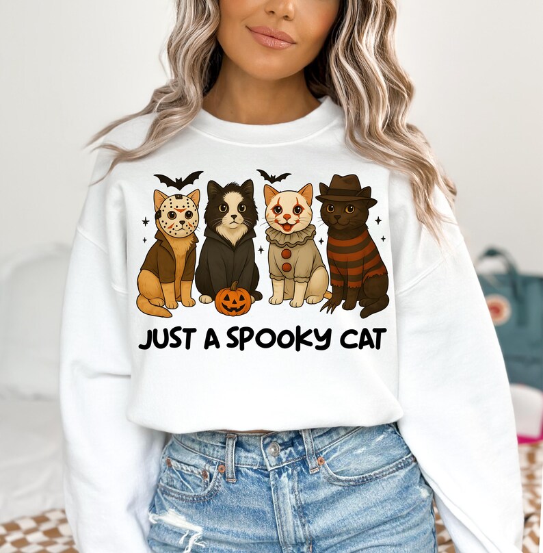 May include: White sweatshirt featuring four cartoon cats dressed in Halloween costumes. The cats include a Jason Voorhees cat, a black cat, a clown cat, and a Freddy Krueger cat. The text "JUST A SPOOKY CAT" is printed below the cats.