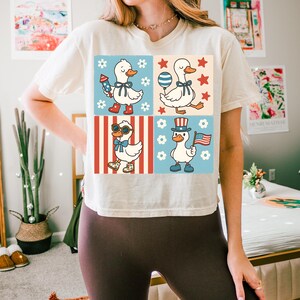 May include: A white t-shirt with a graphic of four ducks in different patriotic outfits. The ducks are wearing red, white, and blue hats, sunglasses, and holding American flags and fireworks. The background is a red, white, and blue checkered pattern.