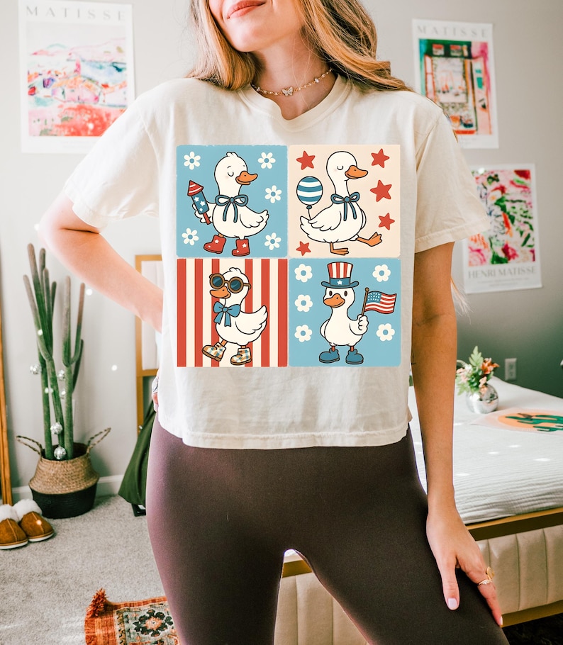 May include: A white t-shirt with a graphic of four ducks in different patriotic outfits. The ducks are wearing red, white, and blue hats, sunglasses, and holding American flags and fireworks. The background is a red, white, and blue checkered pattern.