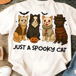 May include: White t-shirt featuring four cartoon cats dressed in Halloween costumes. The cats are dressed as Jason Voorhees, a vampire, a clown, and Freddy Krueger. The text "JUST A SPOOKY CAT" is printed below the cats.