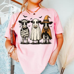 May include: Pink t-shirt featuring three cows dressed in Halloween costumes: a mummy, a ghost, and a witch. The cows are standing on grass with bats flying above them. A fun and festive design for the spooky season.