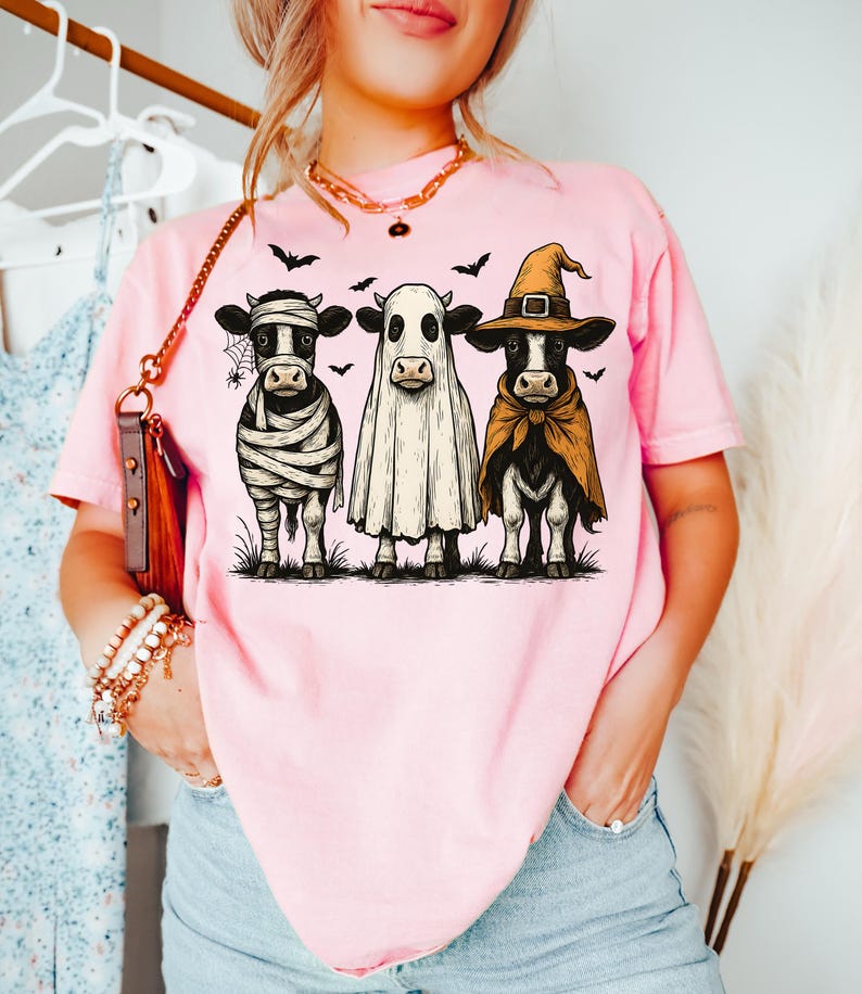May include: Pink t-shirt featuring three cows dressed in Halloween costumes: a mummy, a ghost, and a witch. The cows are standing on grass with bats flying above them. A fun and festive design for the spooky season.