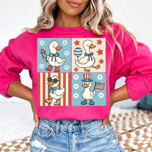 May include: Pink sweatshirt with a graphic of four cartoon ducks wearing patriotic themed outfits. The ducks are wearing red, white, and blue clothing and accessories. The sweatshirt has a crew neck and long sleeves.