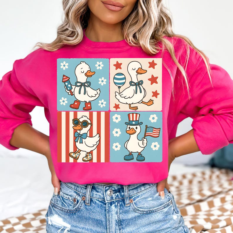 May include: Pink sweatshirt with a graphic of four cartoon ducks wearing patriotic themed outfits. The ducks are wearing red, white, and blue clothing and accessories. The sweatshirt has a crew neck and long sleeves.
