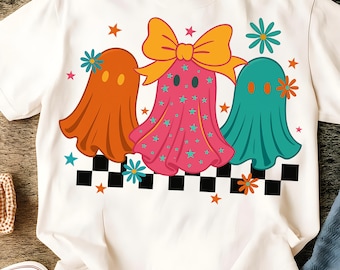 Può includere: Una t-shirt bianca con una stampa grafica di tre fantasmi sopra una striscia a scacchi bianchi e neri. Il fantasma a sinistra è arancione, quello centrale è rosa con stelle verde acqua e un grande fiocco giallo, quello a destra è verde acqua.