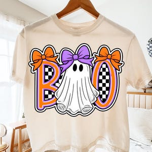 May include: A cream-coloured t-shirt with a Halloween design. The word "BOO" is displayed with orange and purple accents. The "O" is a ghost with a purple bow. The letters have a checkered pattern.