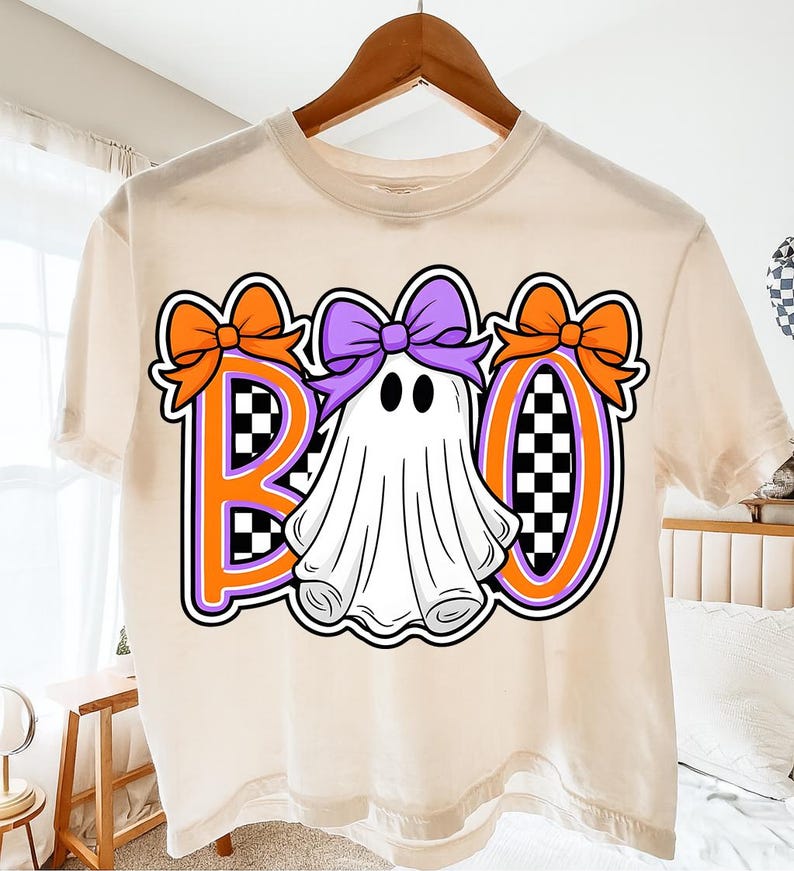 May include: A cream-coloured t-shirt with a Halloween design. The word "BOO" is displayed with orange and purple accents. The "O" is a ghost with a purple bow. The letters have a checkered pattern.