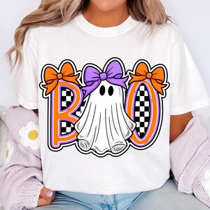 May include: White t-shirt with a Halloween graphic. The word "BOO" is displayed in orange and purple letters, with a ghost in the middle. The letters have a black and white checkered pattern.