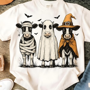 May include: White t-shirt featuring a Halloween-themed graphic. The design showcases three cows dressed as a mummy, a ghost, and a witch, with bats flying above. The cows are standing on grass, creating a festive and whimsical scene.