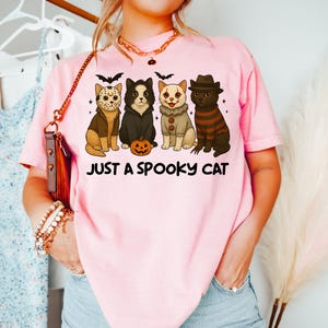 May include: Pink t-shirt with the text "JUST A SPOOKY CAT". The shirt features four cartoon cats dressed in Halloween costumes, including a hockey mask, a black cat, a clown, and a striped sweater. A small jack-o'-lantern is also present.