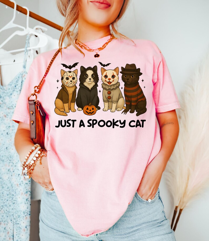 May include: Pink t-shirt with the text "JUST A SPOOKY CAT". The shirt features four cartoon cats dressed in Halloween costumes, including a hockey mask, a black cat, a clown, and a striped sweater. A small jack-o'-lantern is also present.