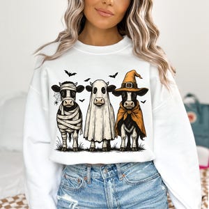 May include: White sweatshirt featuring a Halloween-themed graphic of three cows dressed as a mummy, ghost, and witch. The cows are standing on grass with bats and a spider web in the background. The graphic is in a cartoon style.