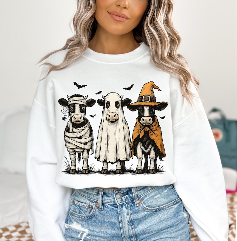 May include: White sweatshirt featuring a Halloween-themed graphic of three cows dressed as a mummy, ghost, and witch. The cows are standing on grass with bats and a spider web in the background. The graphic is in a cartoon style.