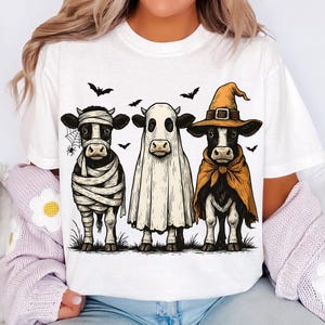 May include: White t-shirt featuring three cows dressed in Halloween costumes. One cow is wrapped in bandages, another is a ghost, and the third is a witch. Bats and a spiderweb are also included in the design.