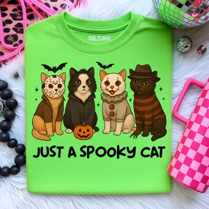 May include: A lime green t-shirt with the text "JUST A SPOOKY CAT." The shirt features four cartoon cats dressed in Halloween costumes: Jason Voorhees, a vampire, a clown, and Freddy Krueger. Bats and stars are also included in the design.