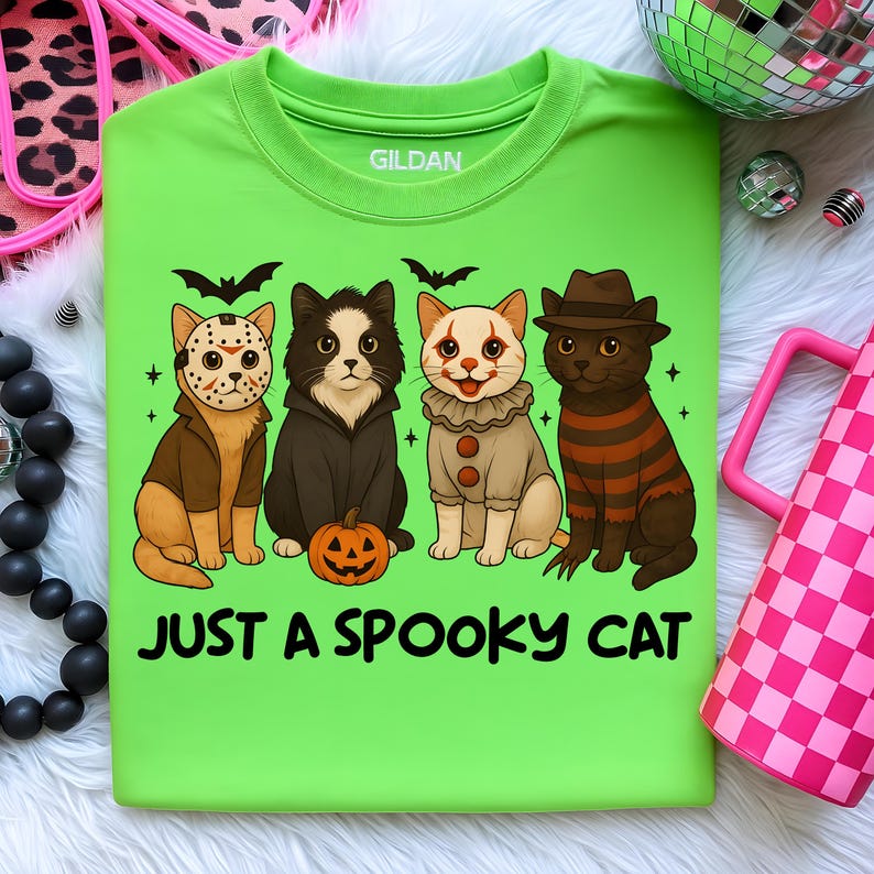 May include: A lime green t-shirt with the text "JUST A SPOOKY CAT." The shirt features four cartoon cats dressed in Halloween costumes: Jason Voorhees, a vampire, a clown, and Freddy Krueger. Bats and stars are also included in the design.