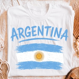 May include: A white short-sleeved t-shirt featuring the word ARGENTINA printed in a light blue brushstroke font. Below the text is a distressed graphic of the Argentine flag, displaying horizontal light blue and white stripes with a yellow sun.