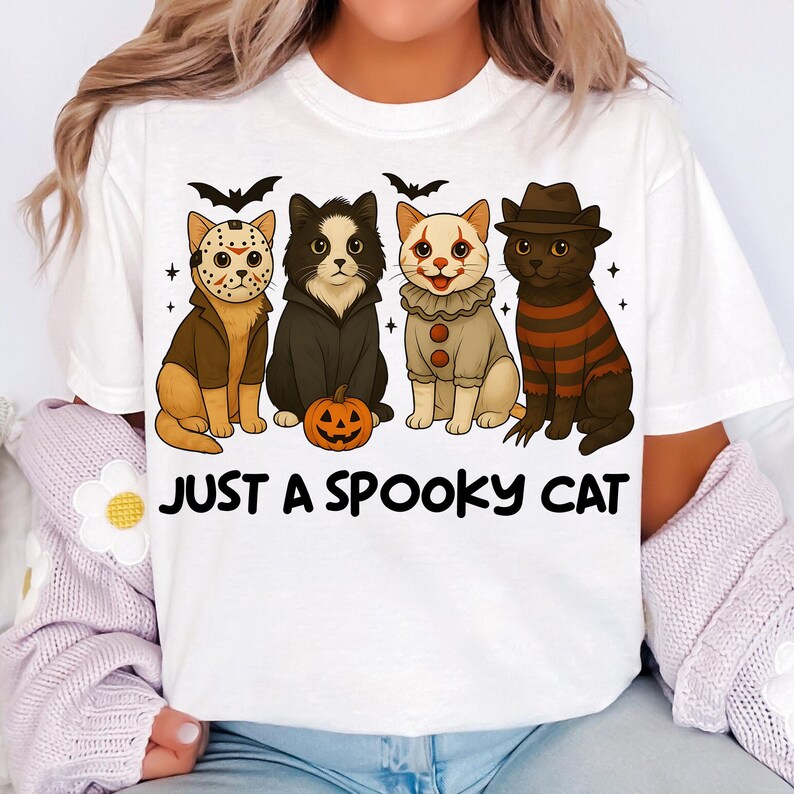 May include: White t-shirt featuring four cartoon cats dressed in Halloween costumes. The cats are depicted as Jason Voorhees, a vampire, a clown, and Freddy Krueger. The text "JUST A SPOOKY CAT" is printed below the cats.