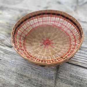 Puede incluir: Una cesta redonda tejida con una base de color beige natural y detalles en rojo y marrón. La cesta tiene un patrón tejido con un diseño de estrella en el centro. La cesta está sobre una superficie de madera desgastada.