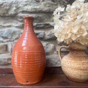 Könnte beinhalten: Zwei Keramikvasen auf einer Holzoberfläche. Die größere Vase ist rot-orangefarben mit schmalem Hals und rundem Körper. Die kleinere Vase ist beige mit Henkel und enthält getrocknete weiße Blumen. Im Hintergrund eine Steinmauer.