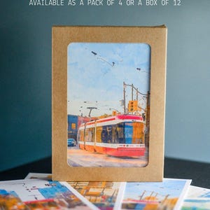 May include: A rectangular brown cardboard box with a window displaying a watercolor print of a red streetcar on city streets. The box features the text AVAILABLE AS A PACK OF 4 OR A BOX OF 12. Several identical prints are spread out below the box.
