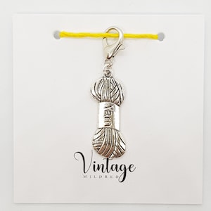 May include: Silver yarn charm with a clip at the top. The charm is shaped like a ball of yarn with the word "yarn" printed on the side. The charm is on a white card with the text "Vintage Mildred" printed on the bottom.