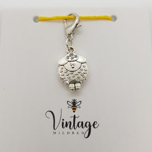 May include: A silver charm featuring a sheep design, attached to a yellow string. The charm has a small loop at the top for attaching to a keychain or other items. The charm is a cute and whimsical addition to any collection.