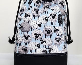 May include: A drawstring bag with a light blue background covered in cartoon sheep. The bag has a black base and a black drawstring closure. The sheep are white with black faces and legs.