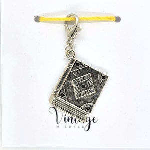 May include: A silver book charm with detailed embossed patterns, attached to a silver clasp and a yellow string. The charm has a vintage aesthetic. The word "Vintage" is printed in cursive below the charm.