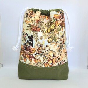 May include: A green drawstring bag with a white cord. The bag is made of fabric and has a print of fairies, leaves, and berries. The bag has a green bottom with a yellow stitch.