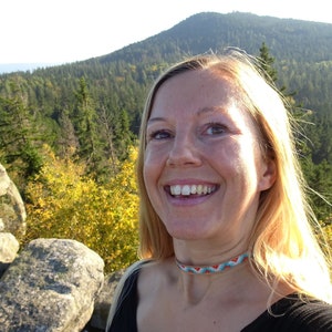 Op de afbeelding: Een vrouw met lang blond haar glimlacht naar de camera terwijl ze op een rotsachtige uitstulping staat. Ze draagt een zwart shirt en een kleurrijke kralenketting. De achtergrond is een weelderig groen bos met een berg in de verte.