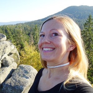 Op de afbeelding: Een vrouw met blond haar glimlacht naar de camera terwijl ze op een rotsachtige uitstulping in een bos staat. De vrouw draagt een zwart shirt en een zilveren choker. Het bos is op de achtergrond, met een bergketen in de verte.