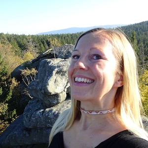 Op de afbeelding: Een vrouw met blond haar glimlacht terwijl ze een zwart shirt en een kralenketting draagt. Ze staat op een rotsachtige uitstulping met een bos op de achtergrond.