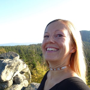 Op de afbeelding: Een vrouw met blond haar lacht terwijl ze op een rotsachtige uitstulping staat. Ze draagt een zwart shirt en een kralenketting. De achtergrond is een bos met een blauwe lucht.
